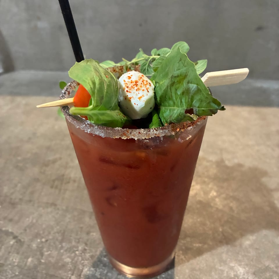Red cocktail with a black straw, salt-and-spice rim, leafy greens, cherry tomato, and mozzarella garnish on a restaurant table.