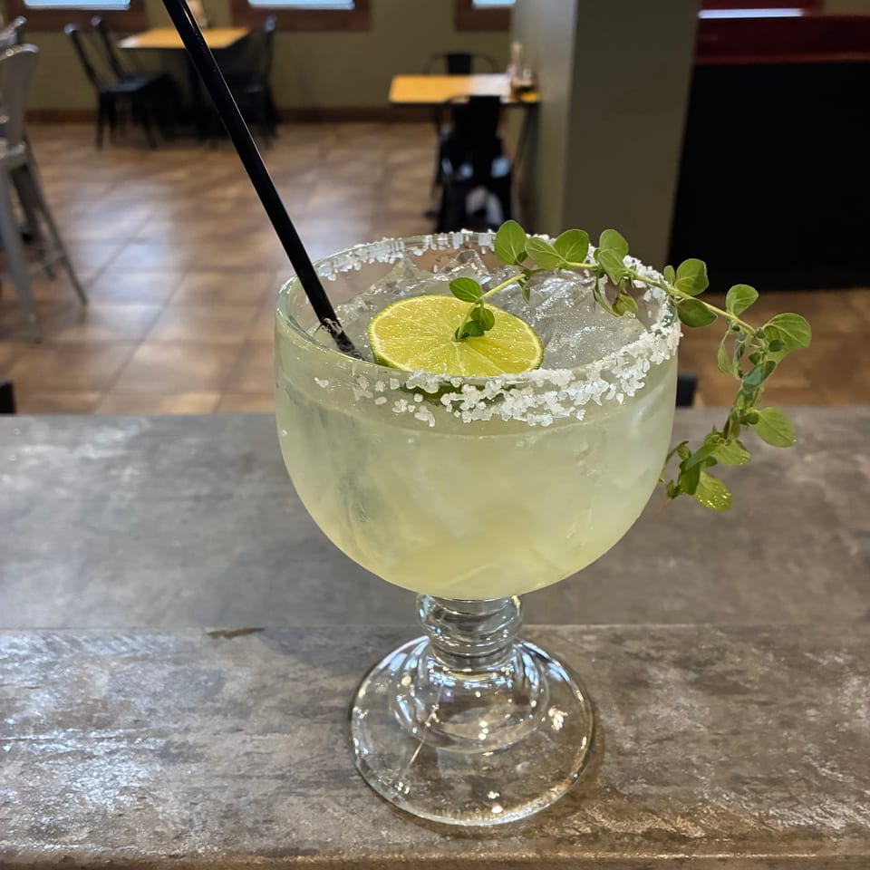 Pale lime cocktail with crushed-ice rim, black straw, lime slices, and fresh herb garnish on a restaurant bar.
