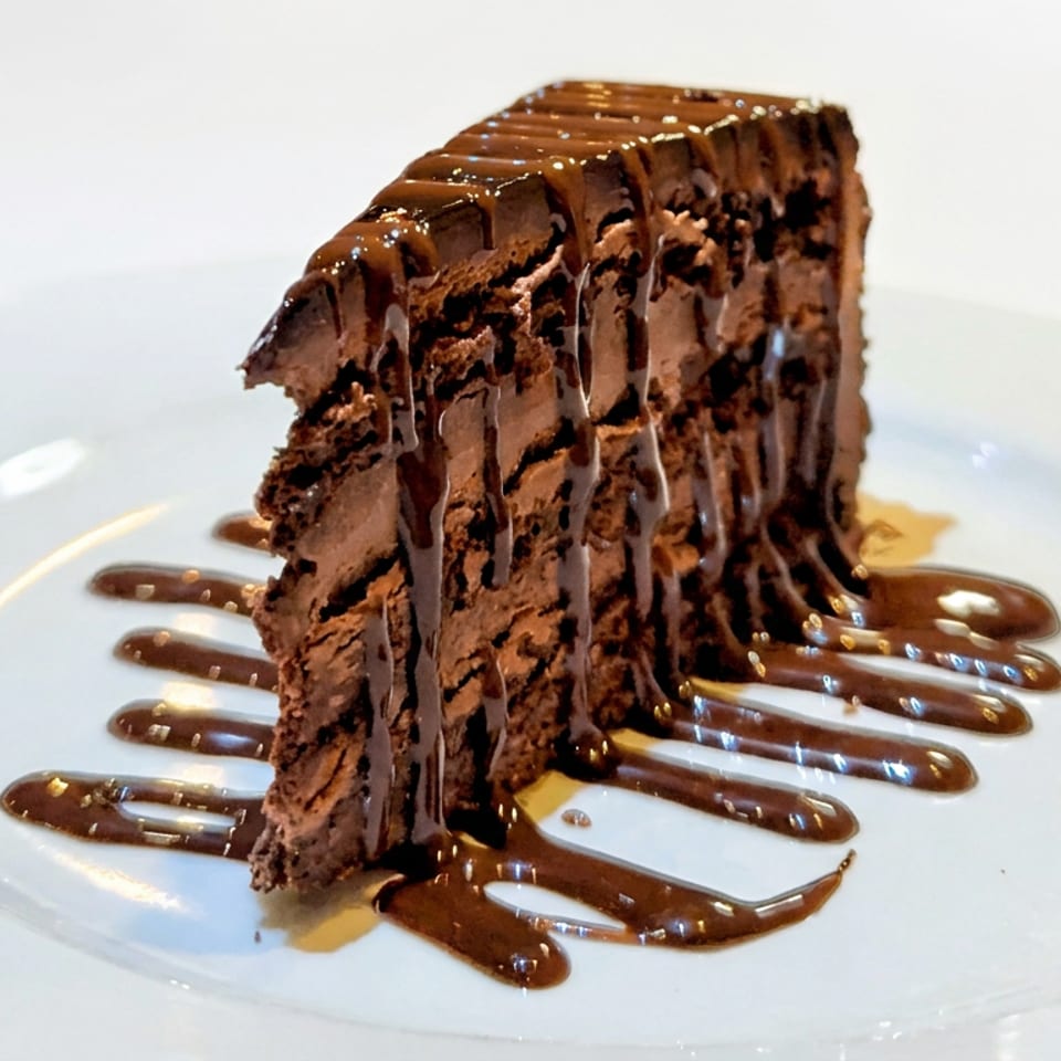 Slice of rich chocolate cake drizzled with chocolate sauce on a white plate.