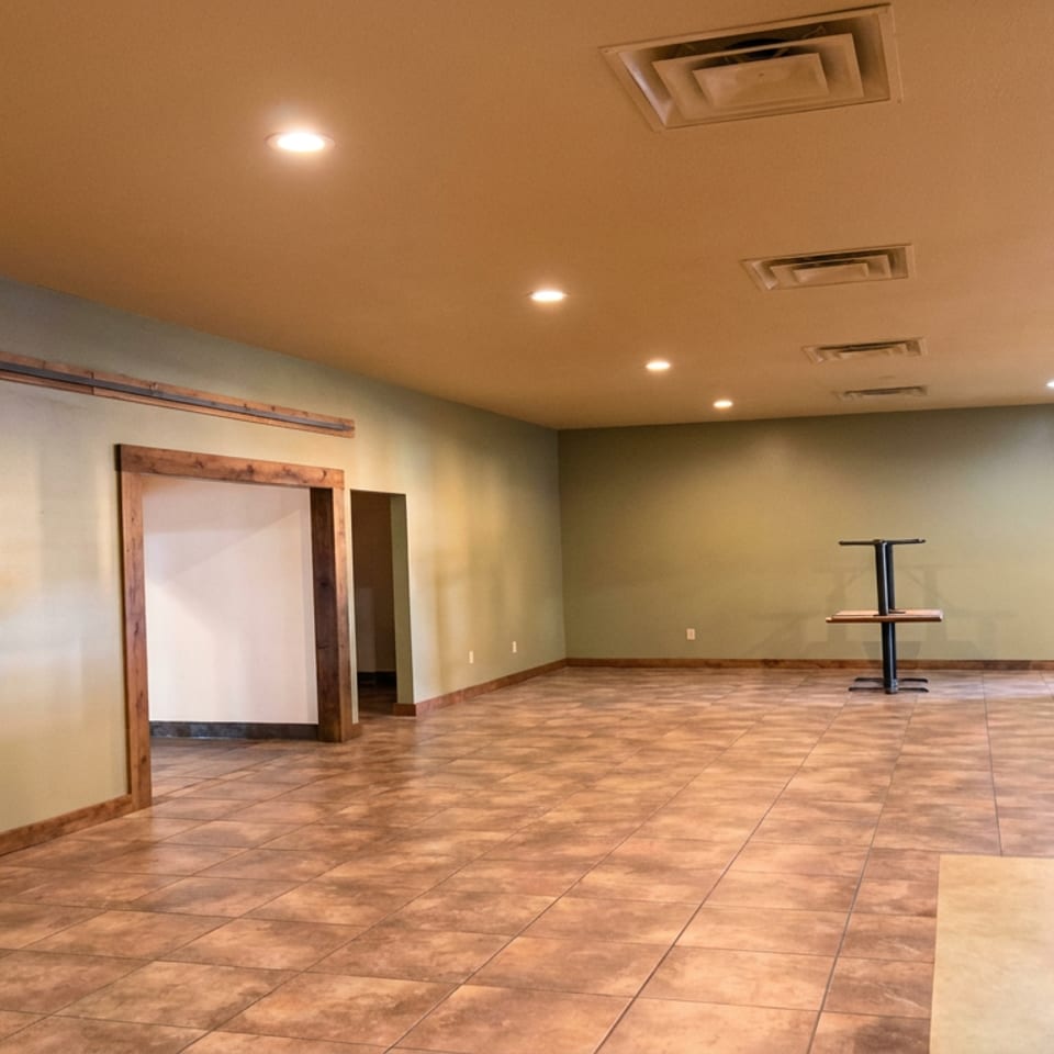 Empty restaurant dining room with tiled floors, warm recessed lighting, wood-trimmed doorway, and a small high-top table.